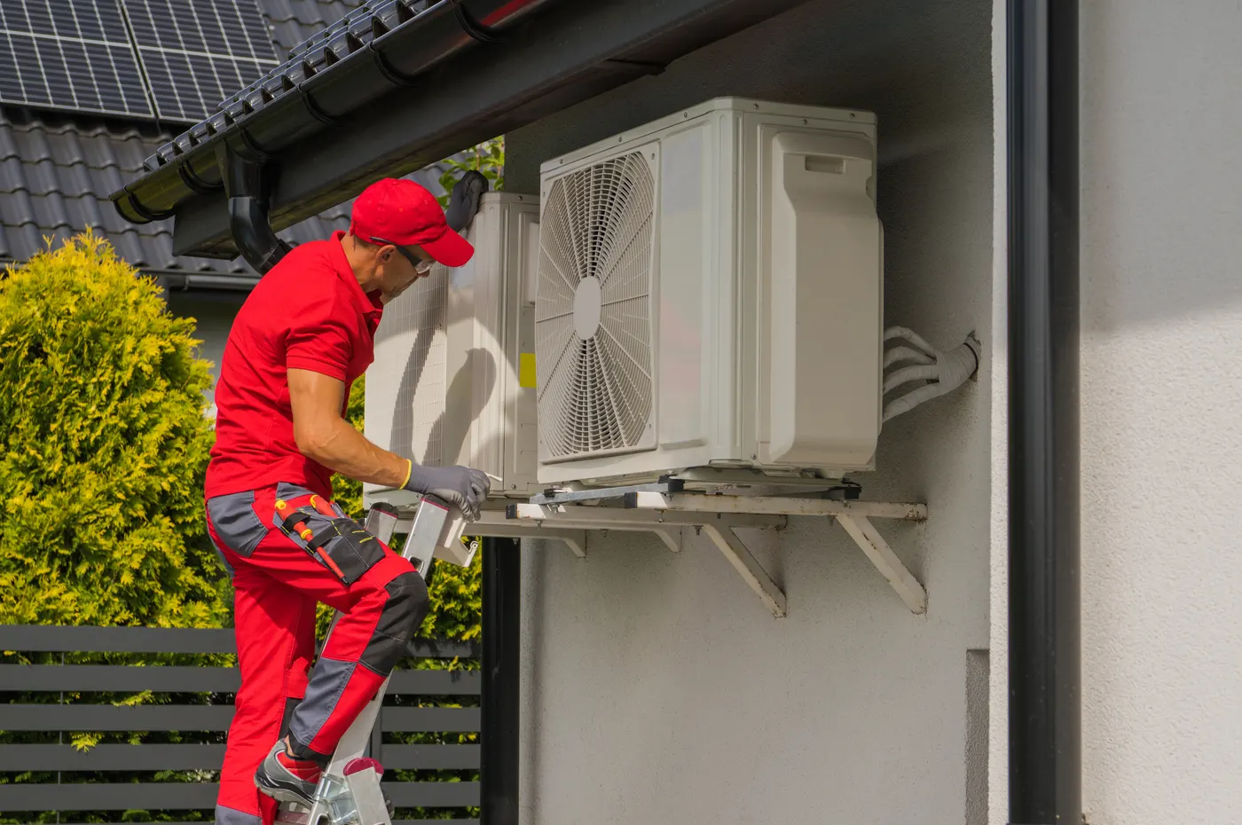 An HVAC technician on a ladder servicing an outdoor heat pump unit