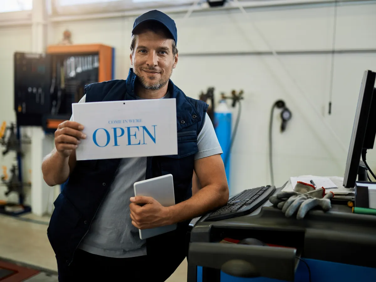 An auto repair shop owner holding an open sign in his workshop