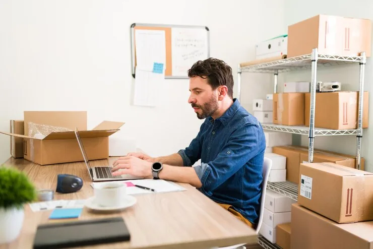 An online store owner typing on a laptop in a stockroom full of shipping boxes